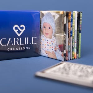 May include: A stack of photo prints with a blue cover featuring the text "CARLILE CREATIONS" and a heart logo. The first photo shows a baby wearing a blue hat. A QR code is in the foreground.