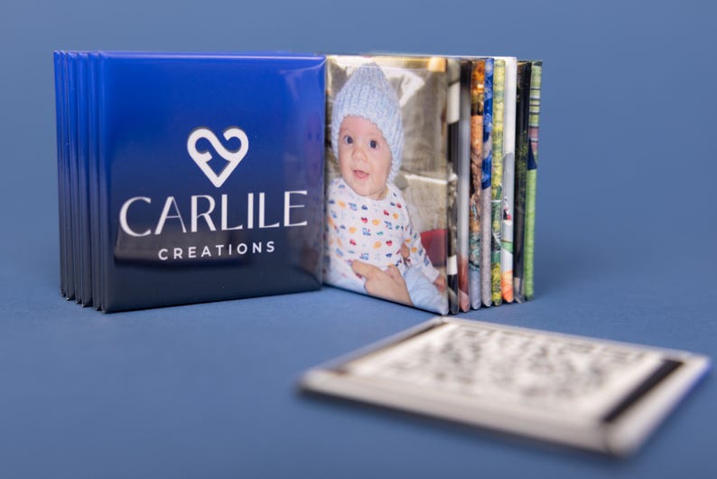 May include: A stack of photo prints with a blue cover featuring the text "CARLILE CREATIONS" and a heart logo. The first photo shows a baby wearing a blue hat. A QR code is in the foreground.