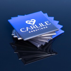 May include: A stack of square, blue and silver business cards. The cards have a gradient from blue to silver and feature the text "CARLILE CREATIONS" in white, along with a white heart-shaped logo. The cards are stacked on a reflective surface.