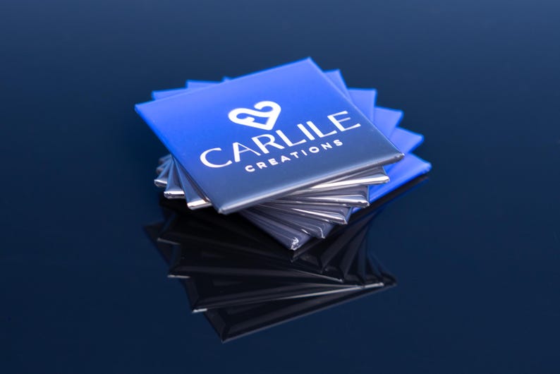 May include: A stack of square, blue and silver business cards. The cards have a gradient from blue to silver and feature the text "CARLILE CREATIONS" in white, along with a white heart-shaped logo. The cards are stacked on a reflective surface.