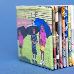 May include: A small, rectangular photo album with a blue background. The cover shows three children with umbrellas on a grassy field. The album's pages are visible on the side, displaying various colorful images.