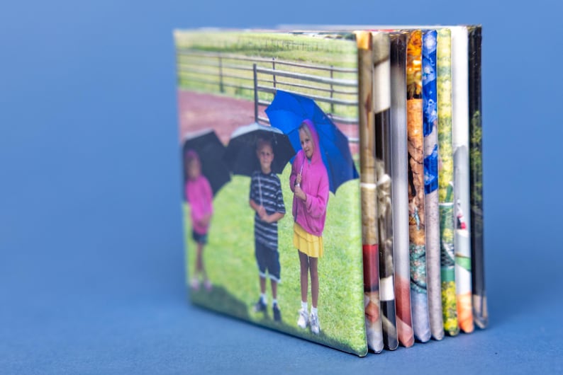 May include: A small, rectangular photo album with a blue background. The cover shows three children with umbrellas on a grassy field. The album's pages are visible on the side, displaying various colorful images.