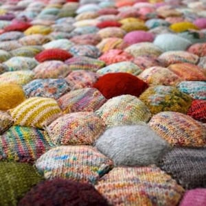 May include: A textile rug composed of numerous small, stuffed, hexagonal-shaped fabric segments. Each individual segment features a unique knit pattern in a variety of colours, including red, yellow, blue, green, grey, and multi-coloured yarn blends.