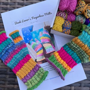 Puede incluir: Un kit de manualidades con mitones sin dedos a rayas multicolores y una caja de ovillos de lana. El kit incluye un patrón titulado "Book Lover's Fingerless Mills" de Jen Yard. Los mitones son principalmente rosas, verdes, amarillos y azules.