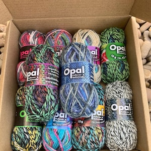Könnte beinhalten: Ein Pappkarton mit zwölf kleinen Knäueln Opal-Wolle in verschiedenen bunten Mustern. Jedes Knäuel hat eine Papierbanderole mit dem Text Opal Pullover- & Sockenwolle, die ein Gewicht von 10g und eine Lauflänge von 42m angibt.