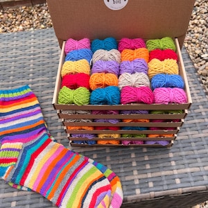 Pode incluir: Uma caixa de papelão cheia de várias camadas de pequenos novelos de lã coloridos em tons de rosa, azul, amarelo, verde, laranja e roxo. Um adesivo redondo na tampa interna diz Thank You. Um par de meias de tricô listradas está ao lado.
