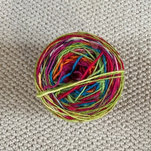 May include: A round ball of yarn featuring a variegated colour palette of lime green, magenta, deep purple, bright orange, and turquoise blue. The fibres are tightly wound in a circular pattern, showcasing a blend of vibrant, multi-coloured strands.