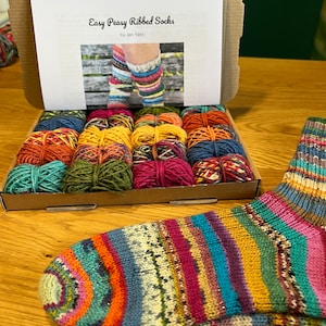 May include: A box of colorful yarn for knitting socks, with a printed instruction booklet titled "Easy Peasy Ribbed Socks" by Jen Yard. The yarn is arranged in rows of different colors, including red, green, blue, and yellow. A finished sock is shown in the foreground, with a colorful striped pattern.