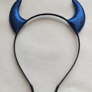 May include: A black headband with two blue devil horns. The horns are a vibrant blue with a textured, shimmering appearance. The headband is thin and black, with rounded tips at the ends.