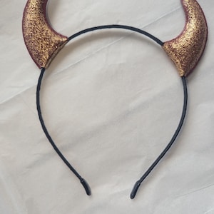 May include: A black headband with two devil horns. The horns are burgundy with gold glitter. The headband is made of a thin, black material.