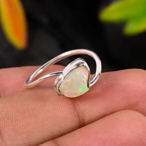 May include: A silver ring featuring a pear-shaped opal gemstone. The opal displays a mix of colors, including white, yellow, and green. The ring's band curves around the finger, creating a modern and elegant design. The gemstone is the focal point.