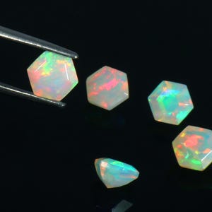 May include: Five loose, hexagonal-cut opals displaying a play-of-color with flashes of red, green, and blue. One opal is held by metal tweezers, while the others are arranged nearby, all featuring a smooth, faceted surface and translucent body.