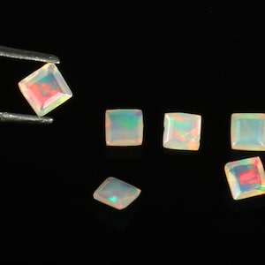 May include: A collection of square-cut opals featuring a translucent white base with iridescent flashes of red, green, and blue. One opal is held by metal tweezers, while five other square opals are arranged on a solid black surface.