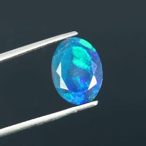 May include: An oval-cut opal gemstone featuring a deep blue base color with vibrant flashes of neon green and electric blue play-of-color throughout the faceted surface, held by a pair of silver-toned metal tweezers against a dark background.