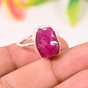 May include: A silver ring featuring a large, raw-cut ruby gemstone. The ruby is a vibrant pinkish-red color and is set in a simple, polished silver band. The gemstone has a rough, natural texture.