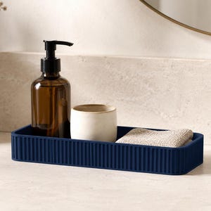 Peut inclure: Un plateau rectangulaire bleu marine aux parois verticales cannelées. À l'intérieur du plateau se trouvent une bouteille en verre brun avec une pompe distributrice noire, une petite tasse en céramique blanc cassé à bord doré et un tissu texturé beige plié.