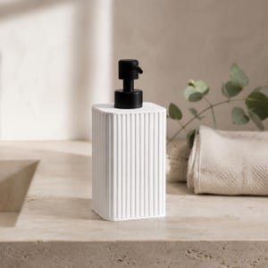 Peut inclure: Un distributeur de savon rectangulaire blanc doté d'une texture nervurée verticale sur les quatre côtés. Le distributeur est surmonté d'un mécanisme de pompe noir mat conçu pour distribuer du savon liquide ou de la lotion par pression.