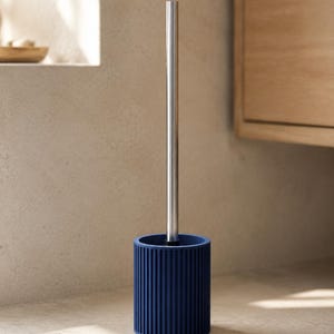 May include: A toilet brush with a long, slender, metallic silver handle and a cylindrical, deep navy blue base featuring a vertical ribbed texture. The brush stands upright, showcasing a clean, functional design with a matte finish on the base.