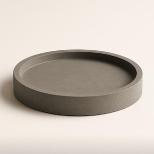 May include: A circular, grey concrete tray with a raised rim. The tray is a solid colour and rests on a white surface. This minimalist tray is suitable for home decor and organisation.