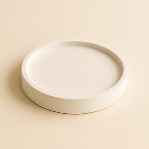 Pode incluir: Uma bandeja redonda de concreto branco sujo com uma borda elevada. A bandeja tem uma superfície lisa e um design minimalista, adequado para guardar pequenos objetos. A cor neutra complementa vários estilos de decoração.