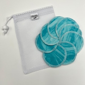 May include: A set of reusable facial rounds in shades of teal and white, with a matching white mesh bag. The rounds have a marbled pattern and are stitched with white thread. The bag has a drawstring closure and a tag that reads "Falling Feathers."