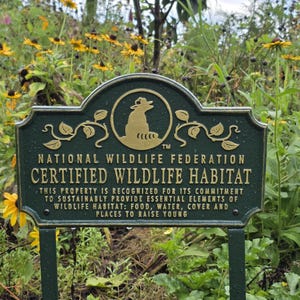 May include: A dark green metal sign with gold lettering, identifying a Certified Wildlife Habitat. The sign features the National Wildlife Federation logo and text detailing the property's commitment to providing essential elements for wildlife.