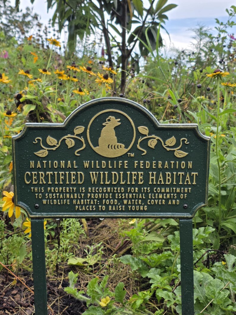 May include: A dark green metal sign with gold lettering, identifying a Certified Wildlife Habitat. The sign features the National Wildlife Federation logo and text detailing the property's commitment to providing essential elements for wildlife.