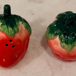 May include: A pair of ceramic salt and pepper shakers shaped like strawberries. Each shaker features a bright red body with yellow seed details and a glossy green stem and leaf top. The shakers have small holes on the upper surface for dispensing.