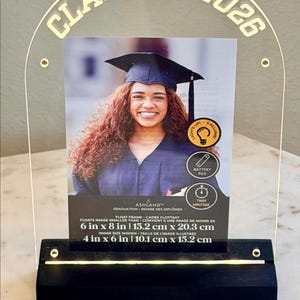 May include: Clear acrylic graduation photo frame with the text "CLASS OF 2026" in gold. The frame holds a 4x6 inch photo, with a black base. The frame has a light-up feature and timer.
