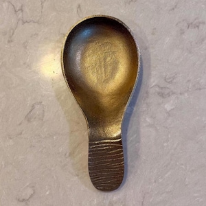 May include: A small, rounded metal scoop with a metallic bronze finish. The bowl of the scoop is smooth and concave, while the handle features a distinct, horizontal ribbed texture across its surface, providing a contrast in the metal finish.