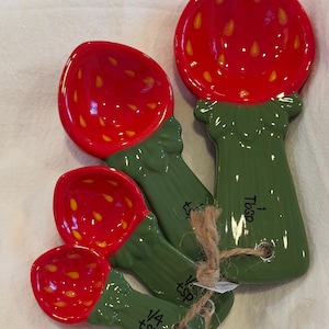 May include: A set of four ceramic measuring spoons shaped like strawberries, featuring red bowls with yellow seed details and green stems as handles. The spoons are labeled 1 Tbsp, 1/2 tsp, 1/4 tsp, and 1/8 tsp, and are tied together with a piece of brown twine.