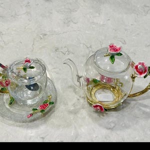 May include: A clear glass teapot and teacup set with gold accents and pink rose embellishments. The teapot has a curved spout and handle, while the teacup sits on a decorative saucer. A spoon rests in the teacup.