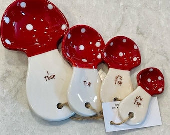 May include: A set of four ceramic measuring spoons shaped like mushrooms with red caps featuring white spots and white stems. The spoons are labeled 1 Tbsp, 1 Tsp, 1/2 Tsp, and 1/4 Tsp in brown text. They are connected by a brown twine string.