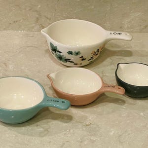 May include: A set of four ceramic measuring cups in various colors. The largest cup is white with floral accents and marked "1 Cup". Other cups are blue, pink, and dark green, labeled "1/2 Cup" and "1/3 Cup".