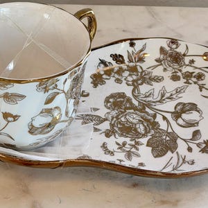 May include: A white ceramic teacup and matching snack plate set featuring a metallic gold floral pattern. The teacup has a gold-toned handle and rim, while the oval-shaped plate has a gold-toned edge. Both pieces are decorated with gold flowers.