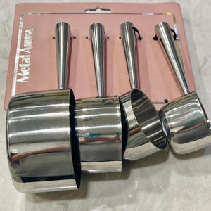 May include: A set of four stainless steel measuring scoops with long handles, displayed on a pink cardboard backing. The scoops are nested together, showcasing their graduated sizes. The text "Metal Aura" is visible on the packaging.