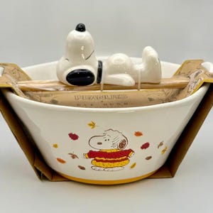 May include: A white ceramic bowl featuring a graphic of Snoopy wearing a yellow and red sweater surrounded by autumn leaves. A ceramic Snoopy figure rests on a wooden spoon across the top, secured by cardboard packaging labeled Peanuts x Rae Dunn.