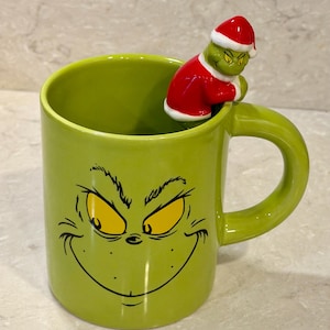 May include: A lime green ceramic mug featuring a printed graphic of a mischievous face with yellow eyes and black outlines. A three-dimensional figurine of a green character wearing a red and white Santa suit is attached to the inner rim.