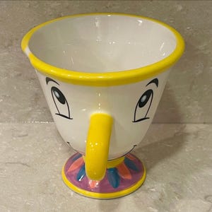 May include: A white ceramic teacup with a yellow rim and handle, featuring a cartoon face with black eyes and a smiling mouth. The base is decorated with a colorful floral design. The cup is approximately 4 inches tall.