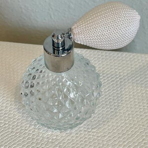 May include: A clear glass perfume bottle with a silver metal top and a white bulb atomizer. The bottle has a textured, diamond-like pattern. The atomizer is pear-shaped and made of a woven white material. The perfume bottle is on a white surface.