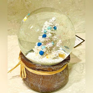 May include: A clear glass snow globe featuring a white Christmas tree decorated with blue ornaments. The globe sits atop a brown fabric base with a raffia bow. Glitter and small fish are inside the globe.