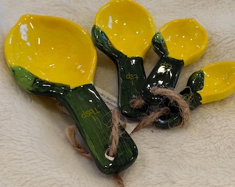 May include: Set of four ceramic measuring spoons. The spoons have a yellow bowl and green handle, resembling lemons. The handles are labeled with measurements in tablespoons and teaspoons. A natural twine string is attached for hanging.