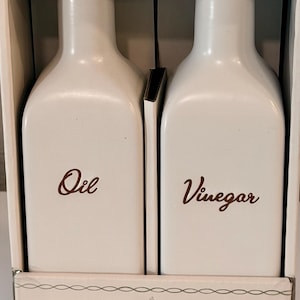 May include: A white oil and vinegar set with gold-colored tops. The bottles are labeled "Oil" and "Vinegar" in a brown script font. The set is packaged in a white box.