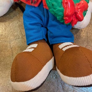 May include: A plush doll wearing blue pants, brown shoes with white stripes, and a red jacket with a plaid collar. The doll is holding a green wreath with a red bow.