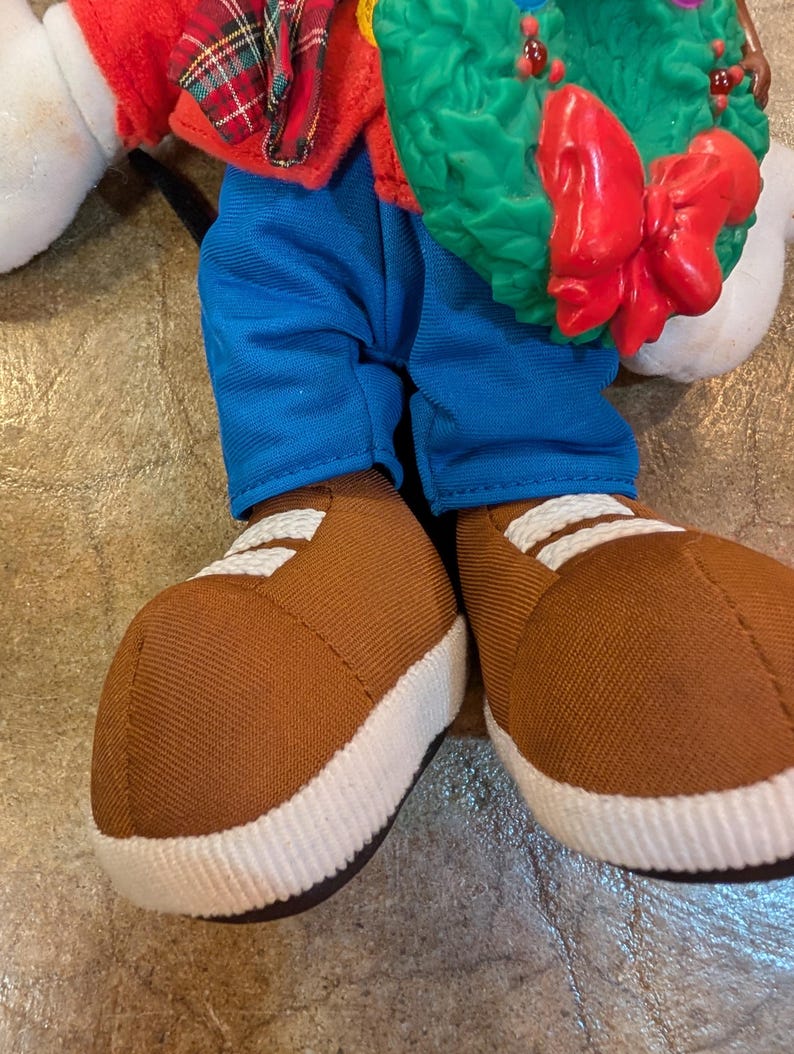 May include: A plush doll wearing blue pants, brown shoes with white stripes, and a red jacket with a plaid collar. The doll is holding a green wreath with a red bow.