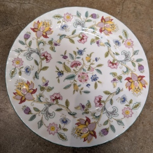 May include: White ceramic plate with a floral design. The plate features a pattern of colorful flowers in shades of pink, yellow, and blue, with green leaves and vines. The rim has a light blue border. A decorative piece for the dining table.