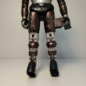 Może przedstawiać: Figurka akcji w stylu vintage przedstawiająca robota w metalicznej srebrno-brązowej kolorystyce. Robot ma czarne dłonie i stopy, ze srebrnymi akcentami na nogach i ramionach. Figurka stoi na białej powierzchni.