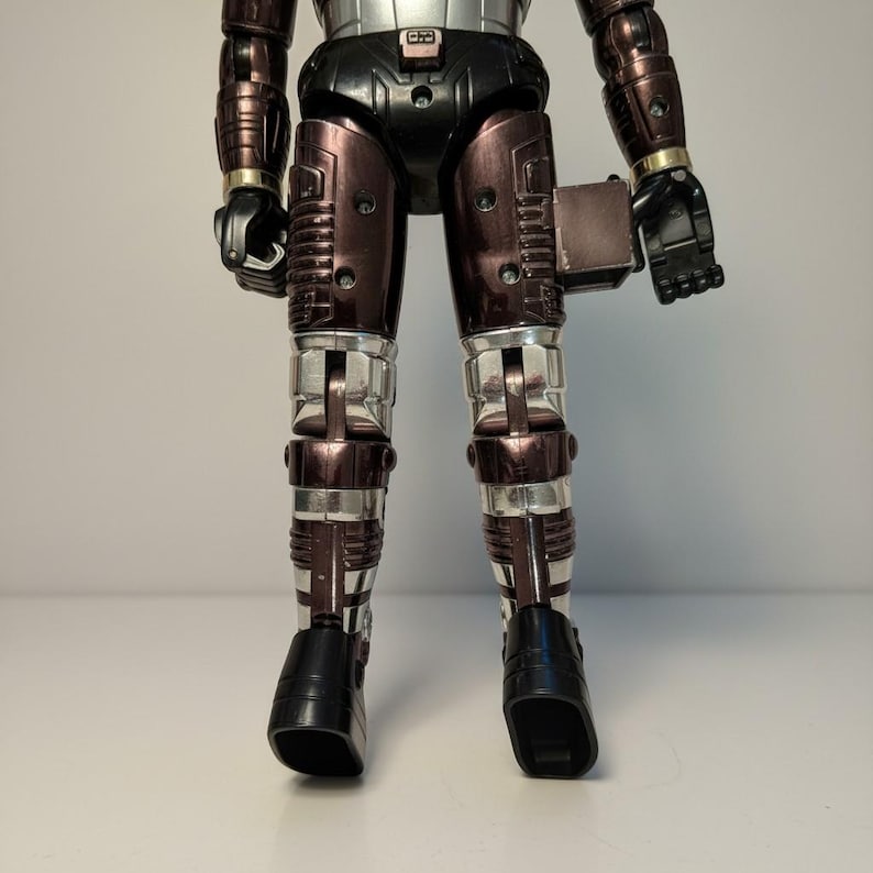 Może przedstawiać: Figurka akcji w stylu vintage przedstawiająca robota w metalicznej srebrno-brązowej kolorystyce. Robot ma czarne dłonie i stopy, ze srebrnymi akcentami na nogach i ramionach. Figurka stoi na białej powierzchni.