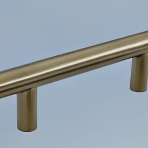 May include: A gold-toned cabinet pull with a cylindrical bar and two cylindrical posts. The hardware is made of metal and has a brushed finish. The pull is approximately 4 inches long.