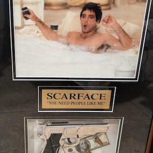 May include: Framed artwork featuring a scene from the movie Scarface. The top portion shows a man in a bathtub. Below, a silver handgun, bullets, and a $100 bill are displayed. The text "SCARFACE" and a quote are also included.